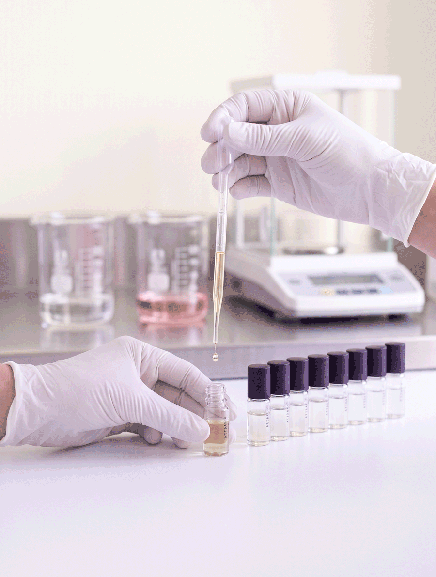 Gloved hands filling a Swiss Emirates attar vial with a pipette, beside a row of capped vials in our facility